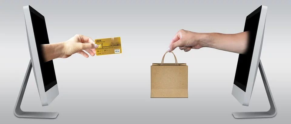 Hands coming out of computer screens facing each other, one holding a credit card and one a paper shopping bag.